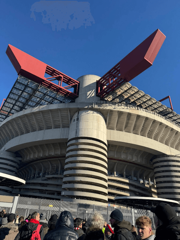 AC Milan San Siro Stadium exterior view showing the iconic concrete cylindrical towers and red steel roof structures with fans gathered outside on matchday