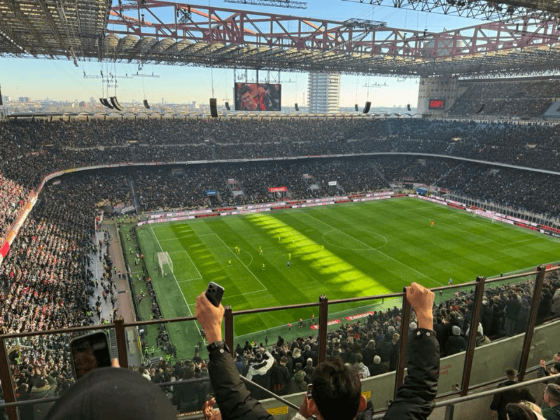 Packed San Siro stadium during an AC Milan home match, showing a full aerial view of the pitch, thousands of fans in the stands, and the iconic multi-tiered stadium structure in Milan