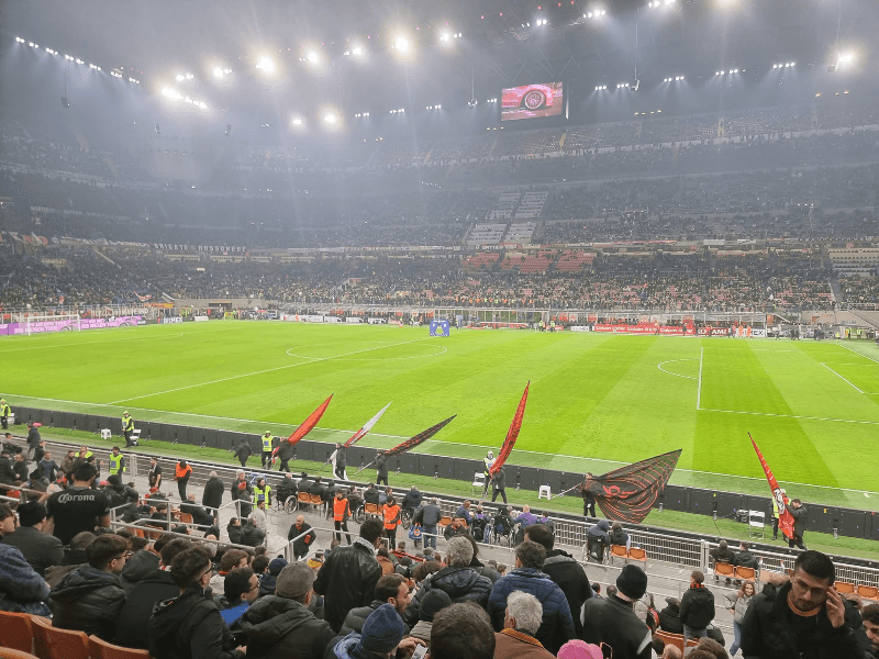 AC Milan matchday atmosphere at San Siro stadium with flag bearers carrying red and black flags along the pitch, surrounded by a packed crowd under bright floodlights