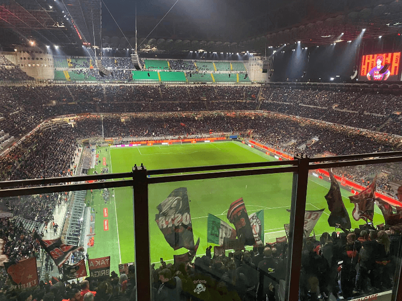 Upper Tier view of San Siro stadium during an AC Milan night match, with fans waving red and black flags in the curva and a packed crowd under floodlights