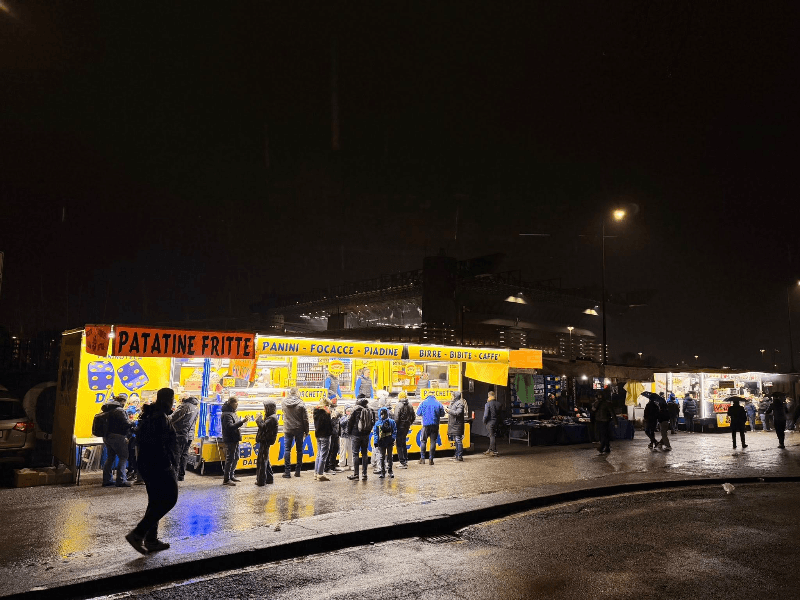 Fans queuing at illuminated Italian street food stalls selling patatine fritte and panini outside San Siro stadium on a rainy night before an AC Milan match