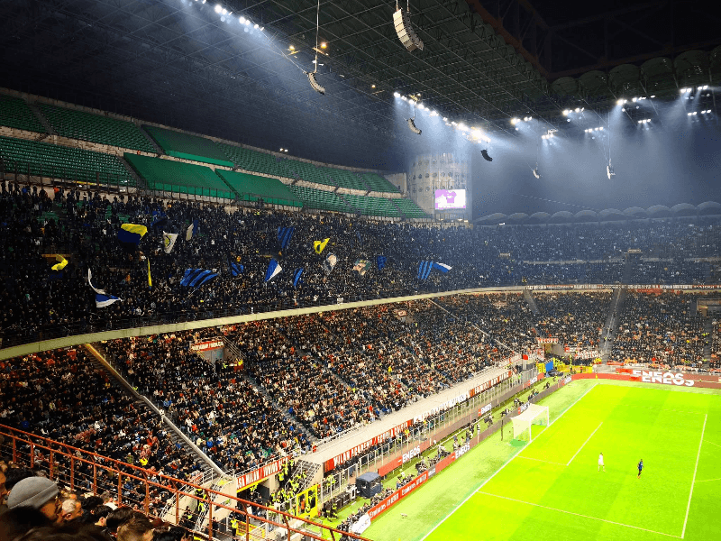 Inter Milan fans in the Curva Nord at San Siro stadium waving blue and black flags during a night match, with the illuminated pitch visible in the foreground